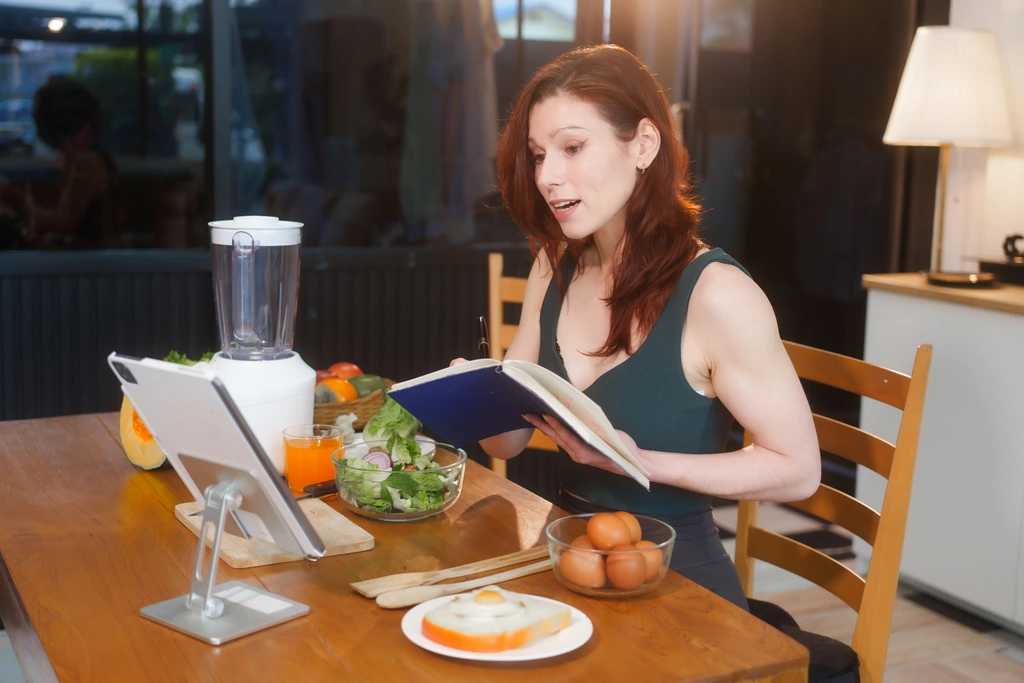 Food blogger Olivia Rosewood sits at her kitchen table developing new recipes for DelishCraze, looking at her notebook with a tablet and fresh ingredients nearby.