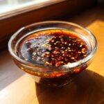 A close-up of a small glass bowl filled with a rich, dark, and flavorful 4-ingredient dumpling sauce with sesame seeds and chili flakes.