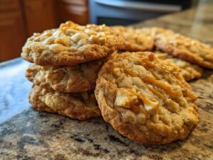 A stack of golden-brown apple pie cookies drizzled with caramel sauce, with more cookies in the background.