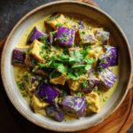 A bowl of delicious aubergine curry, featuring tender purple aubergine pieces in a creamy yellow sauce, garnished with fresh cilantro.