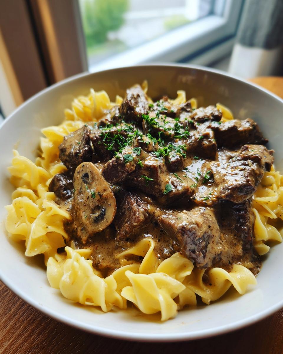 A close-up of a bowl of beef stroganoff with tender beef pieces and mushrooms in a creamy sauce, served over wide egg noodles and garnished with parsley.