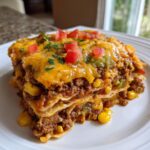 A hearty slice of beef taco casserole on a white plate, layered with seasoned ground beef, corn, cheese, and topped with diced tomatoes.