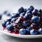 A close-up of glistening, homemade blueberry jam piled high on a white plate, showcasing whole blueberries.