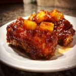 Close-up of glazed brown sugar pineapple chicken pieces on a white plate, glistening with sauce and topped with pineapple chunks.