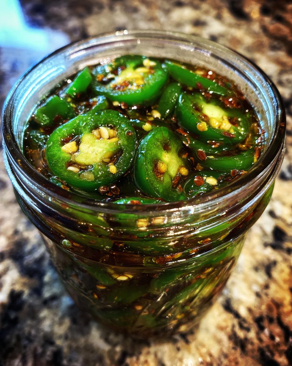 Close-up of sliced candied jalapenos submerged in a dark, sweet brine in a glass jar.