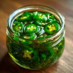 Close-up of a glass jar filled with vibrant green candied jalapenos slices in a syrupy liquid.