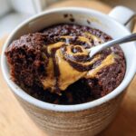 A close-up of a rich chocolate protein mug cake topped with a peanut butter swirl, with a spoonful being taken out.
