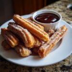 A plate of freshly fried churros coated in cinnamon sugar, served with a side of rich chocolate dipping sauce.