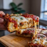 A hand holds a slice of crispy Eggplant Parmesan, showing melted mozzarella cheese and rich tomato sauce.