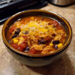 A close-up of a bowl of Crockpot Chicken Enchilada Soup topped with shredded cheese, black beans, corn, and tomatoes.