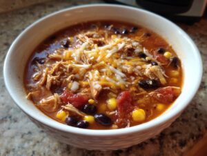 A close-up of a white bowl filled with Crockpot Chicken Enchilada Soup, topped with shredded cheese.