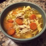 A close-up of a bowl of delicious crockpot chicken noodle soup, featuring shredded chicken, wide egg noodles, and sliced carrots.