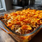 A close-up of a bubbly Dorito Casserole in a glass baking dish, topped with melted cheese and crushed Doritos.