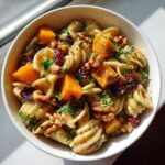 A bowl of fall pasta salad featuring pasta, butternut squash cubes, dried cranberries, walnuts, and parsley.