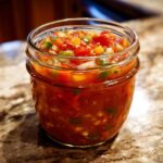 A close-up of a glass jar filled with vibrant fresh tomato canned salsa, showing chopped tomatoes, onions, and jalapeños.
