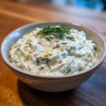 A close-up of creamy Tzatziki sauce in a rustic bowl, garnished with fresh dill and lemon zest.