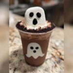 A spooky ghost chocolate milk drink decorated with a marshmallow ghost and cookie crumbs.