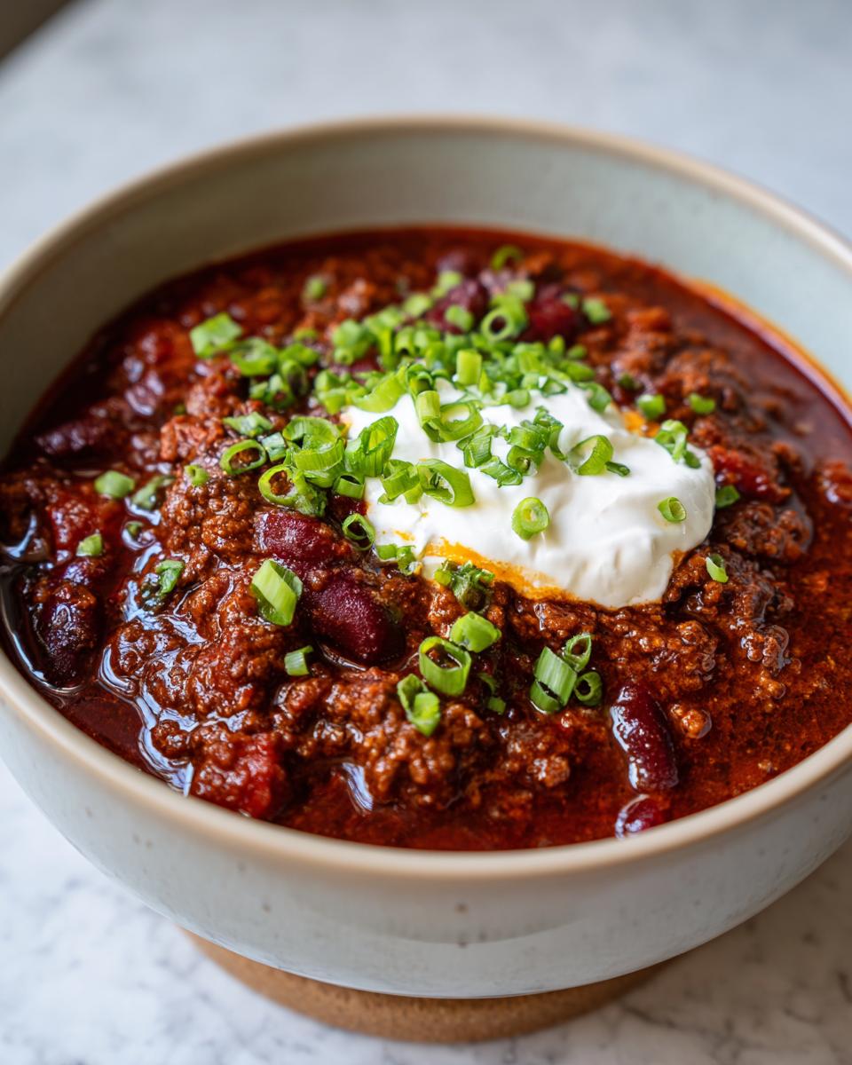 A close-up of a bowl of hearty 1-pot chili topped with a dollop of sour cream and chopped green onions.