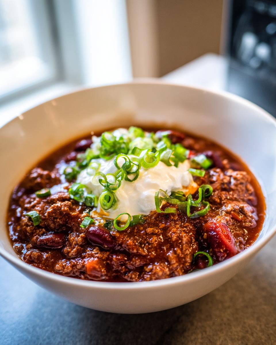 A close-up of a bowl of hearty 1-pot chili topped with sour cream and chopped green onions.
