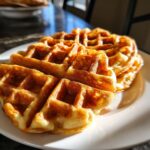 Close-up of golden brown high protein waffles stacked on a white plate, glistening with syrup.