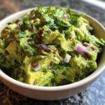 A close-up of a bowl filled with chunky, vibrant green homemade guacamole, garnished with red onion and cilantro.