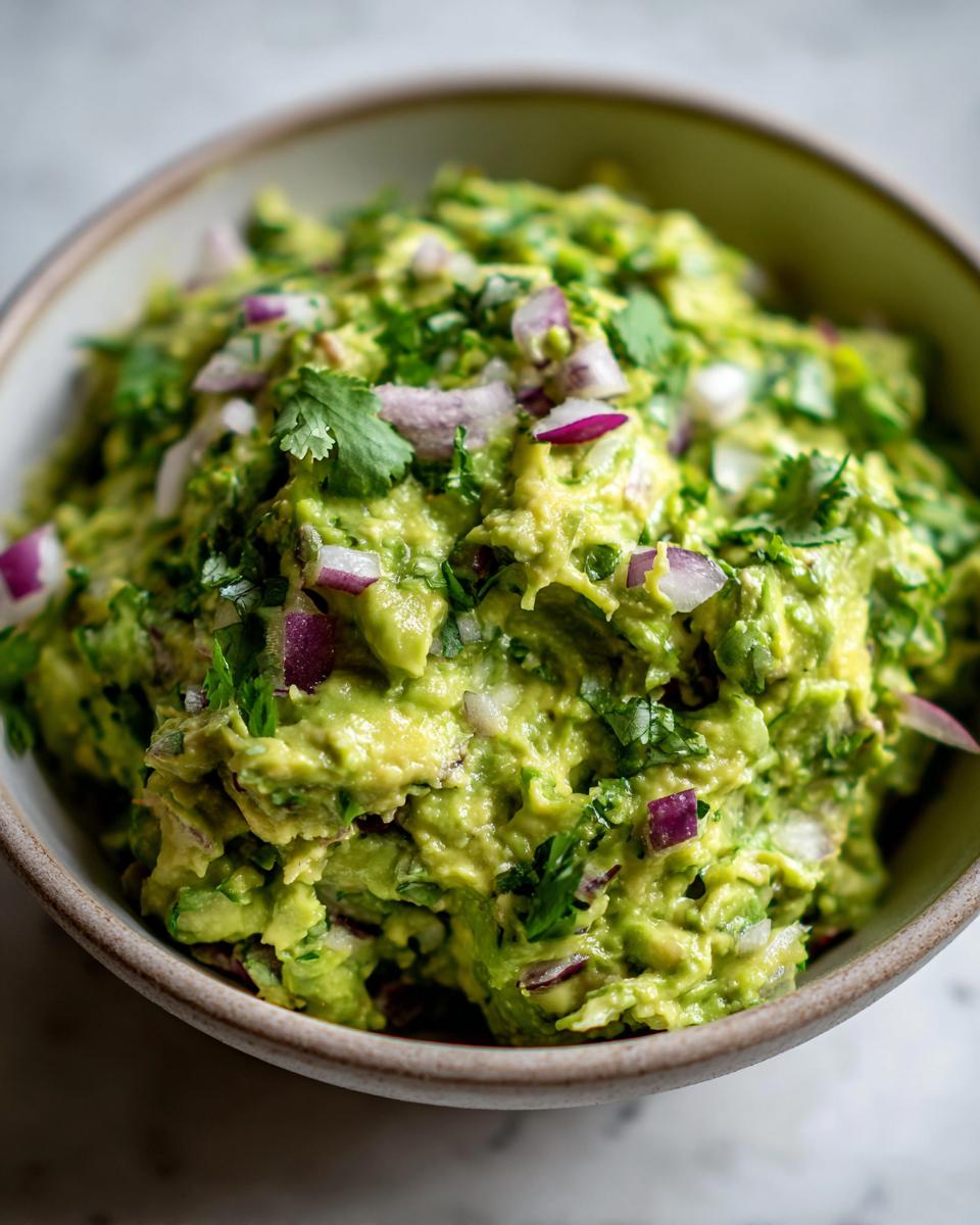 A close-up of a bowl filled with chunky, vibrant green Homemade Guacamole, topped with red onion and cilantro.
