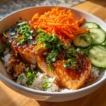 A glistening piece of honey sriracha salmon sits atop white rice in a bowl, with shredded carrots and cucumber slices on the side.