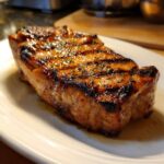 A perfectly grilled pork chop, coated in a rich marinade glaze and sprinkled with herbs, served on a white plate.