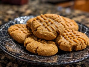 A close-up of several perfect peanut butter cookies with classic criss-cross fork marks, arranged on a decorative glass plate.