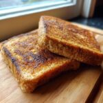Close-up of two golden-brown slices of protein French toast stacked on a wooden cutting board.