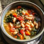 A close-up of a rustic bowl filled with Tuscan white bean soup, featuring white beans, kale, carrots, and tomatoes.