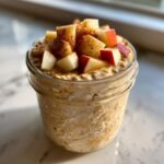 Close-up of apple cinnamon overnight oats topped with diced red apples and cinnamon spice in a glass jar.