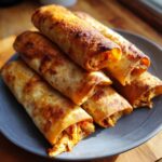 A close-up of a stack of golden-brown baked chicken taquitos on a gray plate, showing shredded chicken filling.