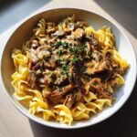 A bowl of creamy beef stroganoff for two served over wide egg noodles, garnished with fresh parsley.