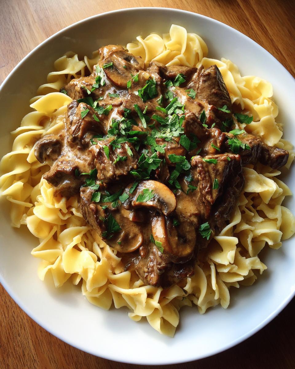 A close-up of beef stroganoff for two served over wide egg noodles, topped with mushrooms and fresh parsley.