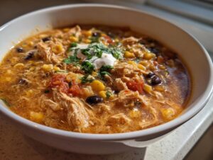 A close-up of a bowl of creamy chicken tortilla soup with shredded chicken, corn, black beans, and a dollop of sour cream.