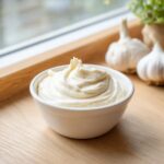 A white bowl filled with thick, creamy garlic sauce, with whole garlic bulbs visible in the background.