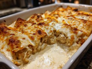 A close-up view of baked creamy queso chicken enchiladas covered in melted, bubbly, golden-brown cheese in a white baking dish.