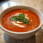 A close-up of a bowl of creamy tomato soup, swirled with cream and topped with fresh basil leaves and black pepper.