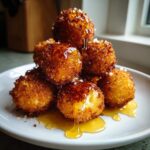 A stack of crispy, golden-brown goat cheese balls drizzled generously with honey on a white plate.