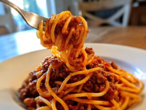 A fork lifting a generous twirl of spaghetti noodles coated in rich meat sauce, ready to eat from a white plate.