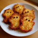 Five golden-brown, crispy, ghost-shaped haunted hash browns decorated with peppercorn eyes on a white plate.