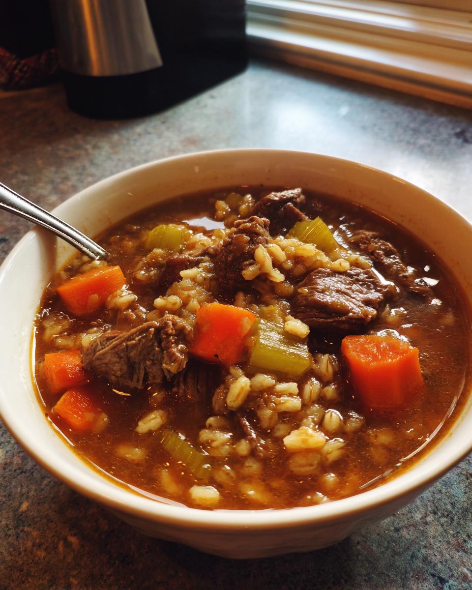 Close-up of a warm bowl of beef barley soup filled with tender beef chunks, barley, bright orange carrots, and celery.