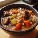 Close-up of a dark bowl filled with rich beef barley soup, featuring chunks of beef, carrots, and celery.