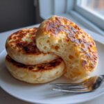 Close-up of three homemade English muffins stacked, one broken open to show the signature nooks and crannies texture.