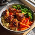 A close-up of a hot honey chicken bowl featuring glazed chicken pieces, quinoa, broccoli, and julienned carrots and peppers.