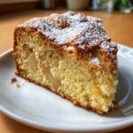 Close-up of a moist slice of Italian apple cake dusted with powdered sugar on a white plate.