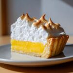 A close-up slice of lemon meringue pie showing bright yellow curd, flaky crust, and perfectly torched meringue peaks.