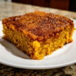Close-up of a square slice of moist, orange pumpkin dump cake with a caramelized top, served on a white plate.