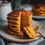 A tall stack of fluffy pumpkin pancakes, with a slice removed and drizzled with syrup.